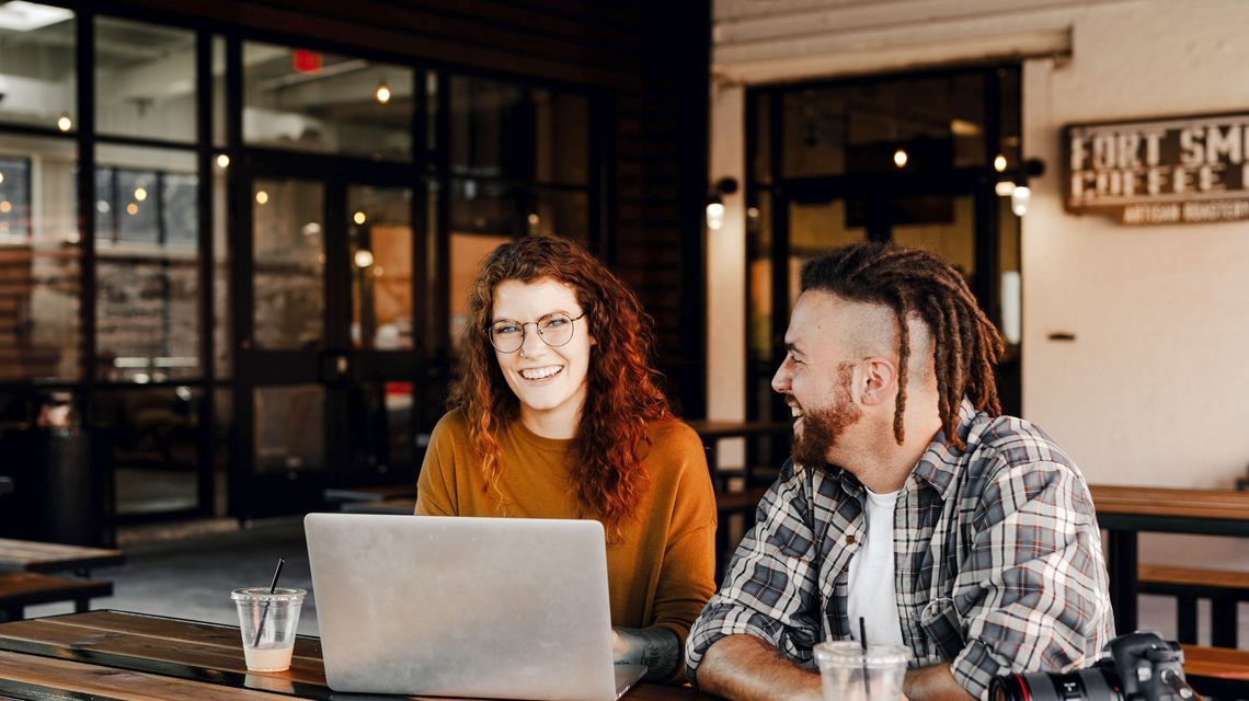 Eine junge Frau und ein junger Mann sitzen zusammen am Tisch und arbeiten an einem Laptop.