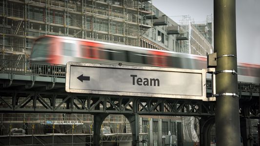 Auf dem Straßenschild steht das Wort "Team". Im Hintergrund fährt eine U-Bahn vorbei.