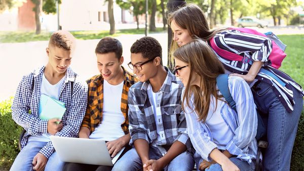 Fünf junge Studierende sitzen draußen auf einer Bank und schauen auf einen Laptop.