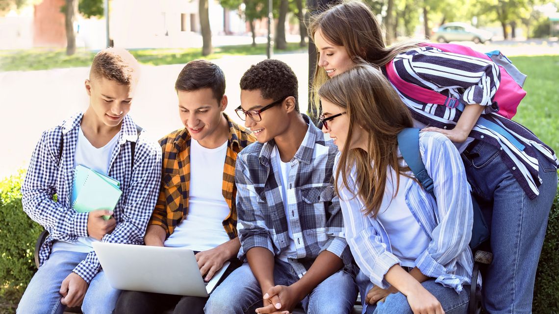 Fünf junge Studierende sitzen draußen auf einer Bank und schauen auf einen Laptop.