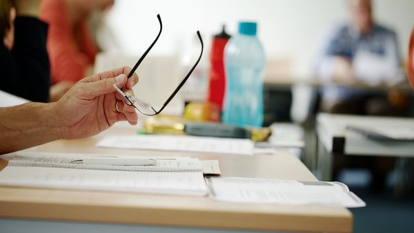 Ein Mann sitzt im Seminar am Tisch, auf dem sein Schreibblock liegt, und hält seine Brille in der Hand.