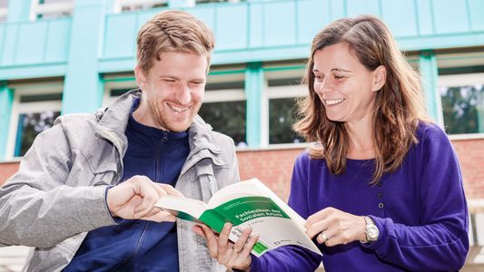 Ein Student und eine Studentin stehen vor der Hochschule und schauen gemeinsam in ein Buch.