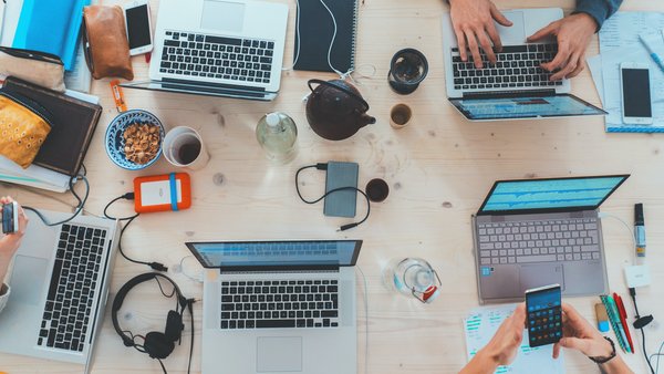 Blick auf den Tisch einer Arbeitsgruppe mit sechs Laptops. Von den Menschen, die am Tisch sitzen, kann man die Hände sehen.