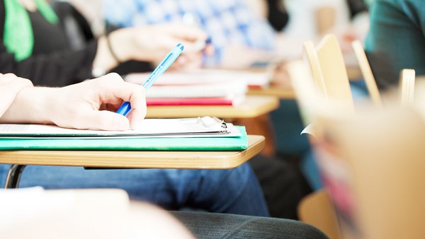 Blick auf einen Klapptisch mit Heften in einem Hörsaal. Studierende im Hintergrund.