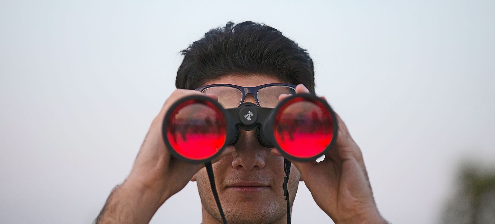 Ein Mann blickt durch ein großes Fernglas, auf den Gläsern spiegelt sich ein roter Himmel.