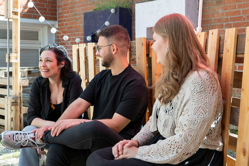 Drei lächelnde Personen sitzen draußen auf Holzmöbeln und unterhalten sich.